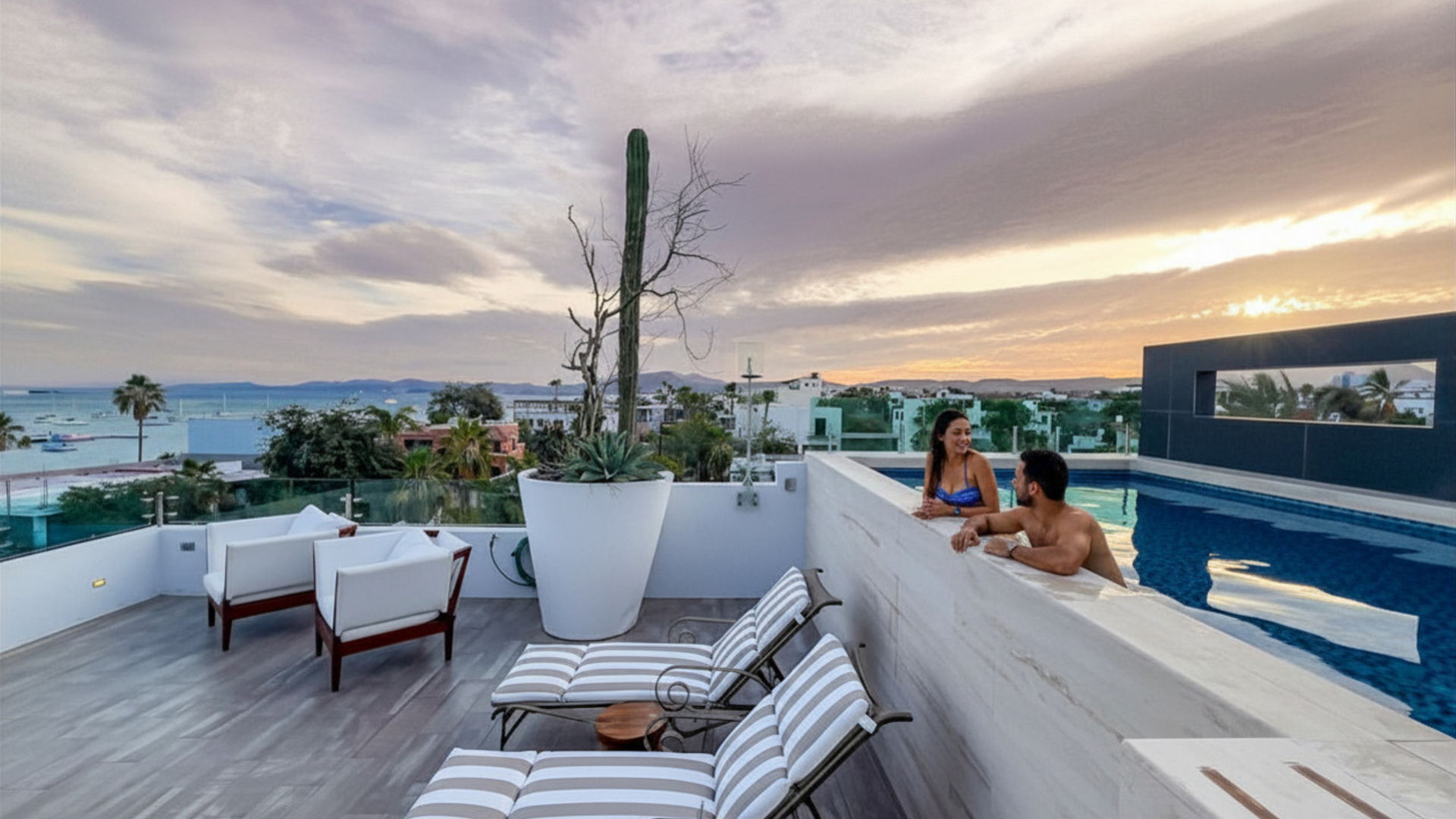 Una pareja en la alberca climatizada del rooftop de Casa Altamar, con la bahía y sus veleros al atardecer
